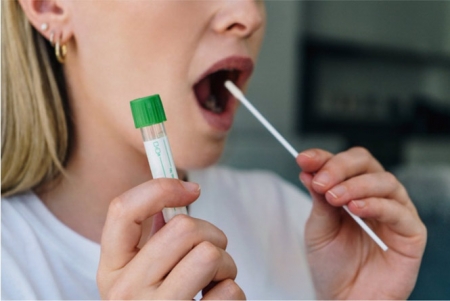 Mujer joven sostiene un hisopo en la boca y sostiene un tubo médico para la prueba de coronavirus (CIRCLE CREATIVE STUDIO/ ISTOCK) 